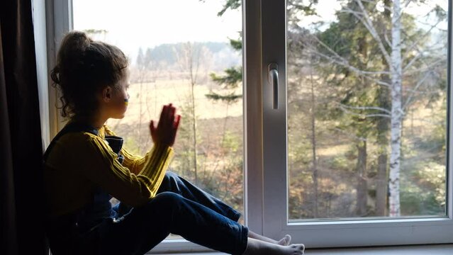 Silhouette of Ukrainian little girl child join hands pray with hope sitting alone on windowsill at home, kid with flag of Ukraine on face. Freedom to Ukraine, Crisis , war concept. Hope for the world