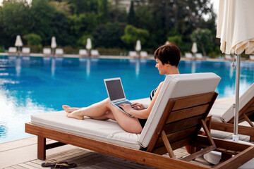a woman in a swimsuit with a laptop lies on a sun lounger by the pool and looks at the monitor...