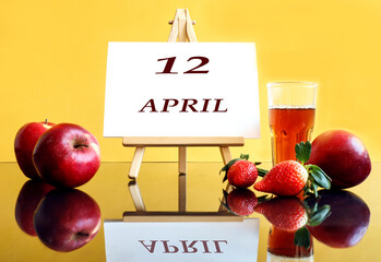 Calendar for April 12: an easel with the inscription 12, april, apples and strawberries next to it, a glass of freshly squeezed juice, yellow background, side view
