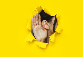 Close-up of a woman's ear and hand through a torn hole in the paper. Bright yellow background, copy space. The concept of eavesdropping, espionage, gossip and tabloids. - obrazy, fototapety, plakaty