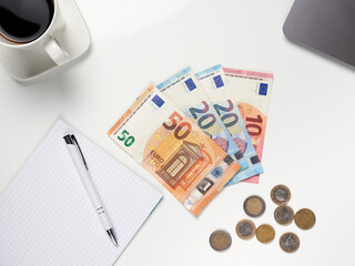 Top view of Euro banknotes and coins lying on the table. Next to it is a cup of coffee, a notebook with a pen and a laptop. White background.