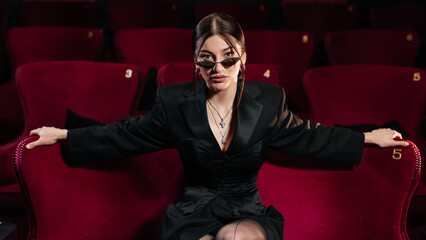 A model wearing all black sitting on a red cinema chair with nice sunglasses looking at camera