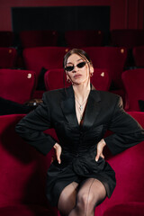 Fototapeta premium A model wearing all black sitting on a red cinema chair with nice sunglasses looking at camera