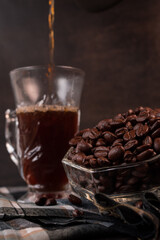 Some black coffee beans in a glass pot next to black coffee cup beind served