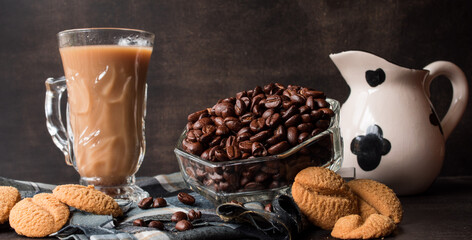 Some black coffee beans  in a glass pot next to cappuccino cup and milk jug