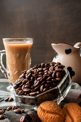 Some black coffee beans  in a glass pot next to cappuccino cup and milk jug