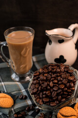 Some black coffee beans  in a glass pot next to cappuccino cup and milk jug