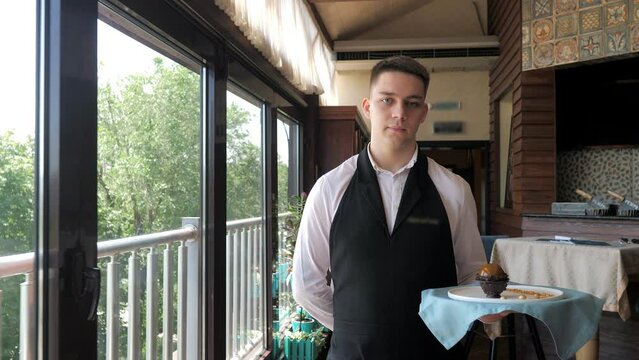 The waiter carries a plate with a delicious dessert along the panoramic windows to the client. A waiter in a white shirt and a black apron, serving on the go, in a modern restaurant. Customer service