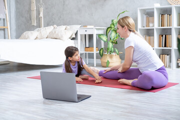 Young mother and little daughter exercising online on fitness mat at home with laptop