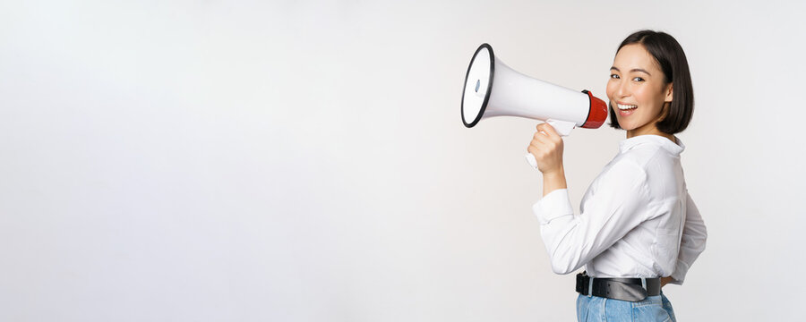 Beautiful Young Asian Woman Talking In Megaphone, Screams In Speakerphone And Smiling, Making Announcement, Shout Out Information, Standing Over White Background