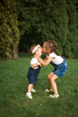 The older girl kisses her little sister on the forehead, both laugh. Tender relationships between brothers and sisters in the family, lack of jealousy. Funny kids having fun in the park on the lawn