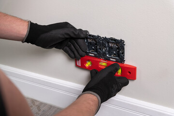An electrician inserts a double grounded electrical outlet into the electrical box in the wall of the room, places the outlet using a spirit level.