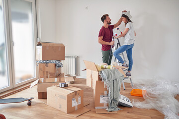 cute couple having fun while preparing to paint white wall in new apartment, standing on a ladder,...