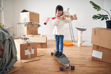 little asian girl smiling, standing on boosted board, holding teddy bear and paint roller, surrounded with unpacked cardboard boxes.