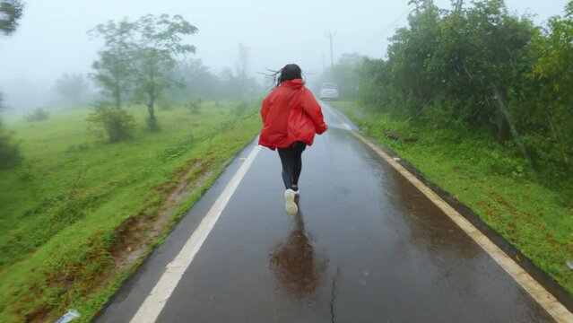 Indian girl wearing red jacket running road at Saputara Hill Station. Gujarat, India. Scenic road covered by green trees on both side during monsoon season.  