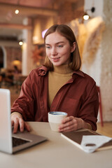 Vertical medium portrait shot of stylish Caucasian woman sitting at cafe table having video call on laptop during coffee break