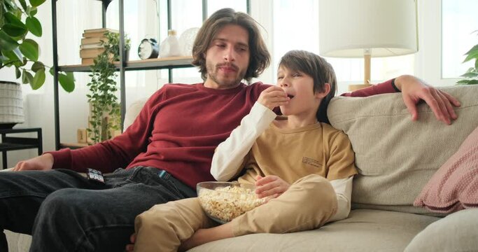 Father and son enjoying watching movie on tv and eating popcorn at home