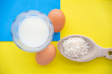 wooden spoon with rice , a glass of milk and two chicken eggs on a blue and yellow surface