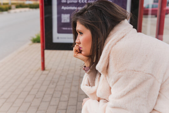 Woman With A Bored Face Waiting For Public Transport At The Bus Stop. Close Shot Of A Young Brunette Girl Discouraged When She Sees That The Bus Does Not Arrive On Time.