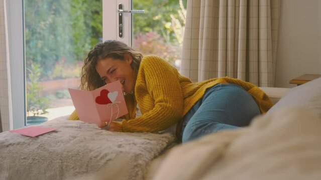 Young woman in cosy warm jumper lying on sofa at home reading romantic valentine's day card - shot in slow motion