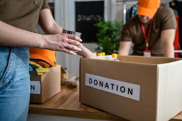 Close up on canned food on the table ready for donation at the charity foundation with unknown caucasian woman volunteer putting stuff in the box