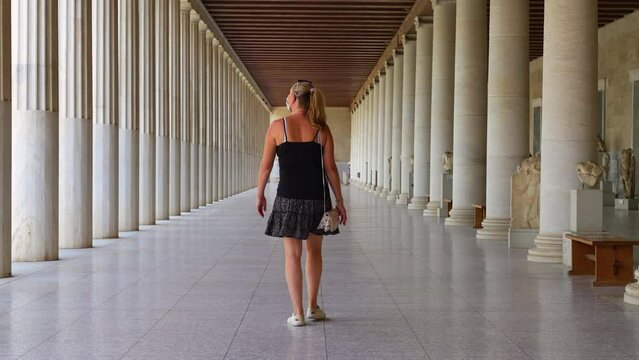 Rearview of female Caucasian tourist slowly walking a long Greek colonnade at the Stoa of Attalos in Athens, Greece