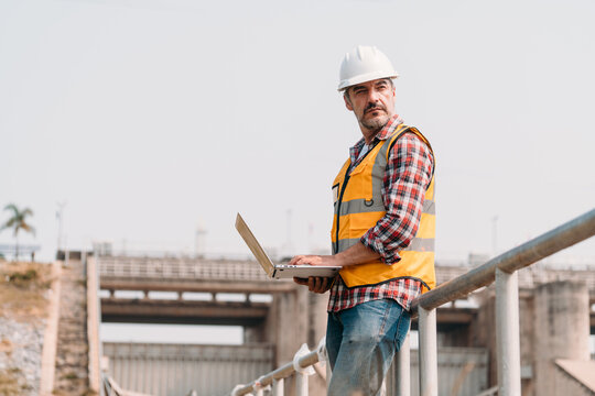 Portrait of power engineer wearing safety jacket and hardhat with laptop computer working at outdoor field site that have water spillway  of hydro power dam electrical generator at the background.
