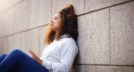 Shes coming up with a tune of her own. Shot of an attractive young woman sitting down and leaning against a building while listening to music in the city.