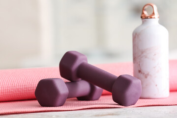 Dumbbells, fitness mat and bottle on table in room, closeup