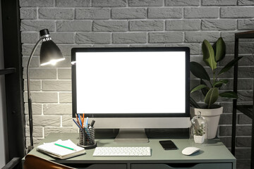 Fototapeta premium Workplace with computer, mobile phone, stationery supplies and glowing lamp near grey brick wall