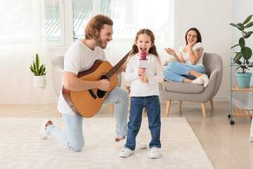 Little girl singing into microphone while her father playing guitar at home