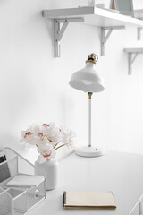 Workplace with modern lamp, notebook and vase with flowers near white wall