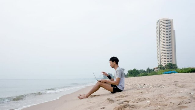 4K 50fps, A handsome Asian man sits at the beach explaining his work to his supervisor, via video call on his laptop computer, comfortably at the beach,work from home.
