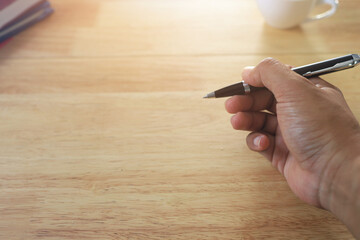 Entrepreneur hand holding a pen on a wooden desk with a white coffee cup. light shines in from the front copy space.