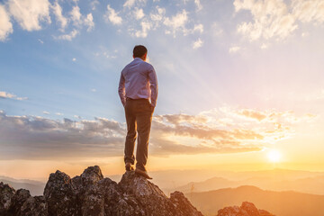 Successful businessman in business suit standing and looking sunset on top rock mountain...