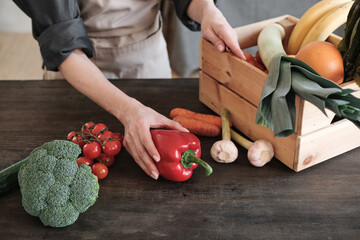 Close-up of unrecognizable woman in apron standing at wooden table and unpacking box of vegetables and fruits