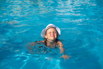 Child learns swim in pool. Happy little girl in white panama hat takes swimming lessons, rejoices at her success and squints from bright summer sun. 