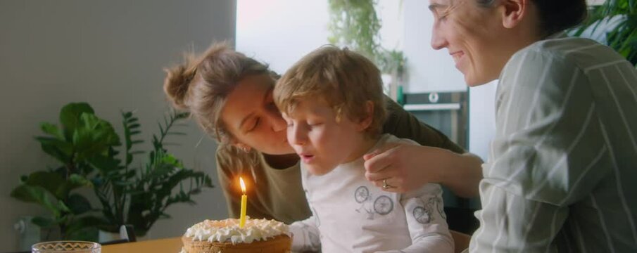Happy LGBTQ family celebrating birthday on kid at home surprised little son blowing candle on cake as loving lesbian mothers greeting and kissing him