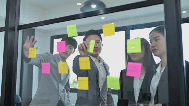 Group of Asian business people tracking tasks on kanban board. Using task control of agile development methodology. Businessman and businesswoman attaching sticky note to scrum task board in office