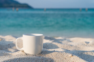 Obraz na płótnie Canvas White coffee cup on white sand beach with blue sea background in morning. Coffee relaxation time in travel holiday concept.
