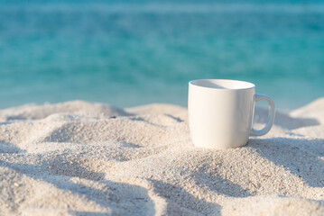 White coffee cup on white sand beach with blue sea background in morning. Coffee relaxation time in travel holiday concept.