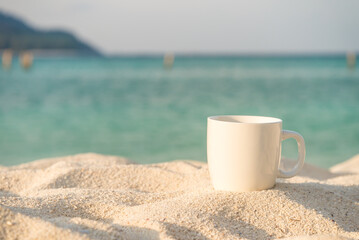 Fototapeta na wymiar White coffee cup on white sand beach with blue sea background in morning. Coffee relaxation time in travel holiday concept.