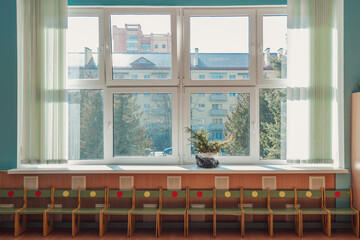 Wooden chairs standing in row under large open window in empty class room at sunny day