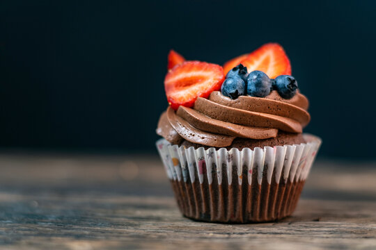 Fresh chocolate mafin on wooden table. Cake is decorated with chocolate cream, blueberries and strawberry slices. Close-up