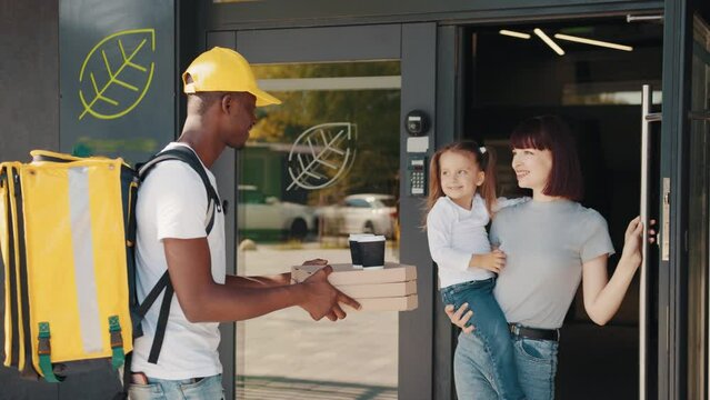 The young courier delivers the parcel directly to the customer's home. Courier concept, door to door delivery. An African-American courier with a thermal bag delivers food to a woman with a child.