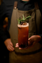 Close-up view of glass of signature cocktail decorated with rosemary on male bartender hand