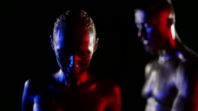 portrait of a couple with golden metallic skin on a black background. the woman lowered head. the man is seen out of focus. red, blue and white light. the dark key. close-up