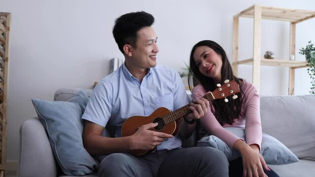 Young Asian couple playing ukulele and sing a song together on sofa in living room at condominium