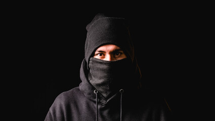 Thief in black balaclava and black cap looking at camera while standing against dark background.