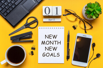 Office workplace with coffee cup, eyeglasses, smart phone, laptop and plant. Date 01 April on wooden block calendar. Text NEW MONTH NEW GOALS on white notepad page. Table desk top view, flat lay